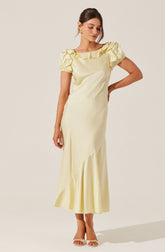 A woman is wearing a long, cream-colored satin dress with short puffed sleeves. The dress features a ruffled neckline and an asymmetrical hemline with a subtle pleated detail near the bottom. She is also wearing gold earrings and nude high-heeled sandals.